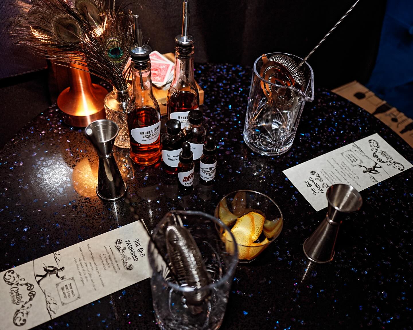 Sparkling black bar table set for mixology: crystal mixing glasses with strainer and bar spoon, metal jiggers, amber syrup bottles, small bitters droppers, bowl of orange twists, peacock feather accent and Old Fashioned cocktail menus.