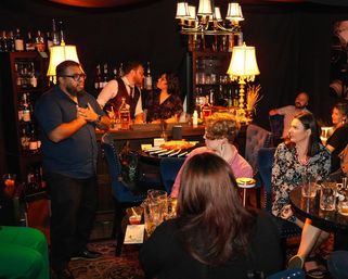 Cozy dimly lit cocktail lounge with a presenter speaking to seated guests, bartenders behind a stocked bar, warm lamps and plush chairs creating a speakeasy vibe.