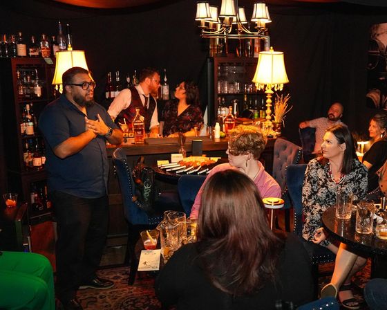 Cozy dimly lit cocktail lounge with a presenter speaking to seated guests, bartenders behind a stocked bar, warm lamps and plush chairs creating a speakeasy vibe.