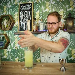 Tattooed bartender mixing a pale green cocktail in a tall glass at a tropical-themed home bar with leafy wallpaper and liquor bottles on a shelf