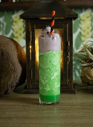 Halloween-themed green slushy mocktail in a tall glass topped with purple foam and black sprinkles, orange-and-black striped straw, rustic lantern backdrop on a wooden table.