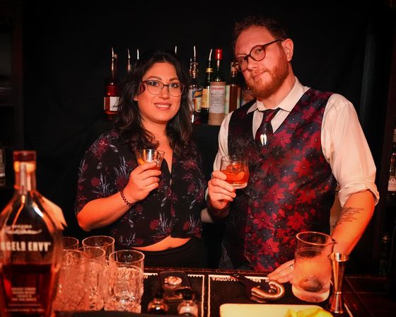 Two bartenders smiling and holding craft cocktails behind a dimly lit speakeasy-style bar with whiskey bottles and glassware