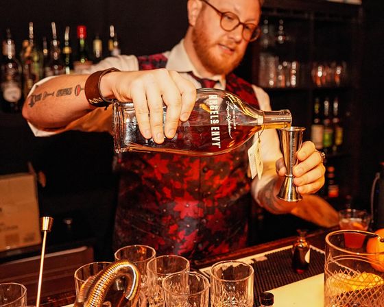 Mixologist in a red patterned vest pours a bottle labeled "Rebel's Envy" whiskey into a stainless steel jigger at a dim cocktail bar, surrounded by glassware and bottles.