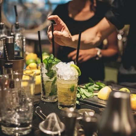 Bartender preparing two zesty mojito-style cocktails with crushed ice, lime wedges, fresh mint and black straws on a busy bar counter.