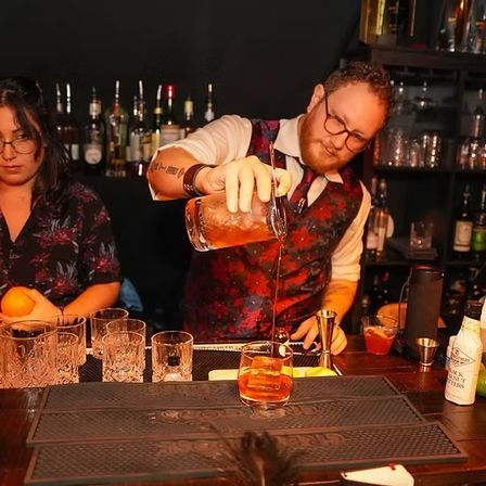 Mixologist in a patterned vest pouring an amber craft cocktail into a rocks glass at a dim craft cocktail bar with bottles and glassware on the counter.
