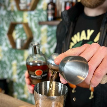 Close-up of hands pouring amber whiskey from a double jigger into a stainless steel cocktail shaker, bottle and tropical-pattern bar shelves blurred in the background.