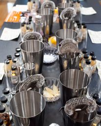 Overhead view of a mixology class table with stainless steel cocktail shakers, hawthorne strainers, jiggers, small bitters bottles, citrus wedges and tasting notes on a black tablecloth.