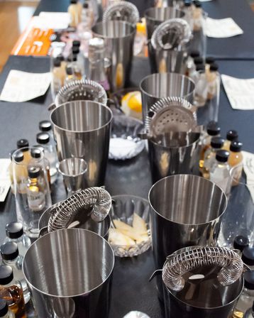 Overhead view of a mixology class table with stainless steel cocktail shakers, hawthorne strainers, jiggers, small bitters bottles, citrus wedges and tasting notes on a black tablecloth.