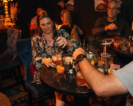 Woman stirring a cocktail at a dimly lit cozy cocktail lounge table with friends, orange-garnished drinks and velvet chairs — urban nightlife bar scene.