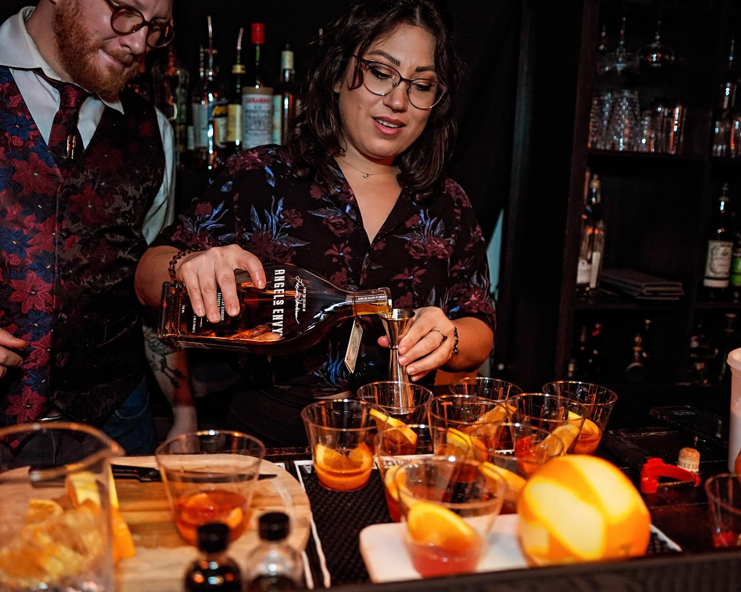 Bartender pouring whiskey from a bottle into a jigger to prepare multiple orange‑garnished cocktails at a dim cocktail bar.