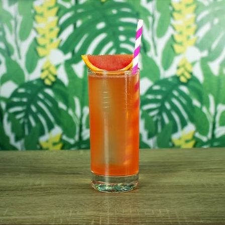 Tall orange tropical cocktail in a highball glass with a pink-striped paper straw and grapefruit wedge garnish on a wooden table against green palm-print wallpaper