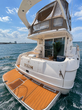 Rear view of a white motor yacht with teak swim platform and enclosed flybridge cruising on blue coastal waters under a sunny, partly‑cloudy sky
