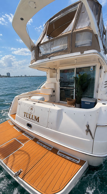 Rear view of a white motor yacht with teak swim platform and enclosed flybridge cruising on blue coastal waters under a sunny, partly‑cloudy sky
