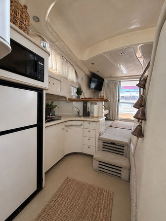Bright beige yacht interior with compact galley—microwave, fridge, sink and curved cabinetry—carpeted steps up to the cockpit and a sliding door revealing docked boats.