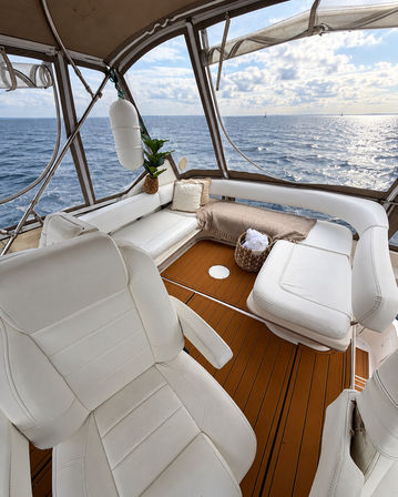 Sunlit yacht cockpit with white leather seating and teak deck, cozy throw and towel basket, panoramic open-ocean view with distant sailboats.