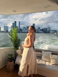 Woman in a beige sundress holding a champagne glass on a yacht deck with potted palm, wake in the water and the Miami skyline under dramatic sunset clouds