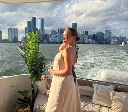 Woman in a beige sundress holding a champagne glass on a yacht deck with potted palm, wake in the water and the Miami skyline under dramatic sunset clouds