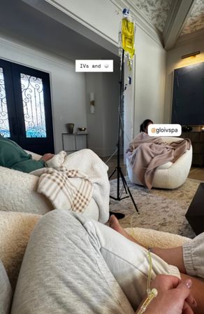 Cozy living room scene with people lounging in plush boucle chairs and blankets while connected to a yellow IV infusion bag on a standing pole — soft neutral decor, patterned throw, and relaxed at-home wellness vibe.