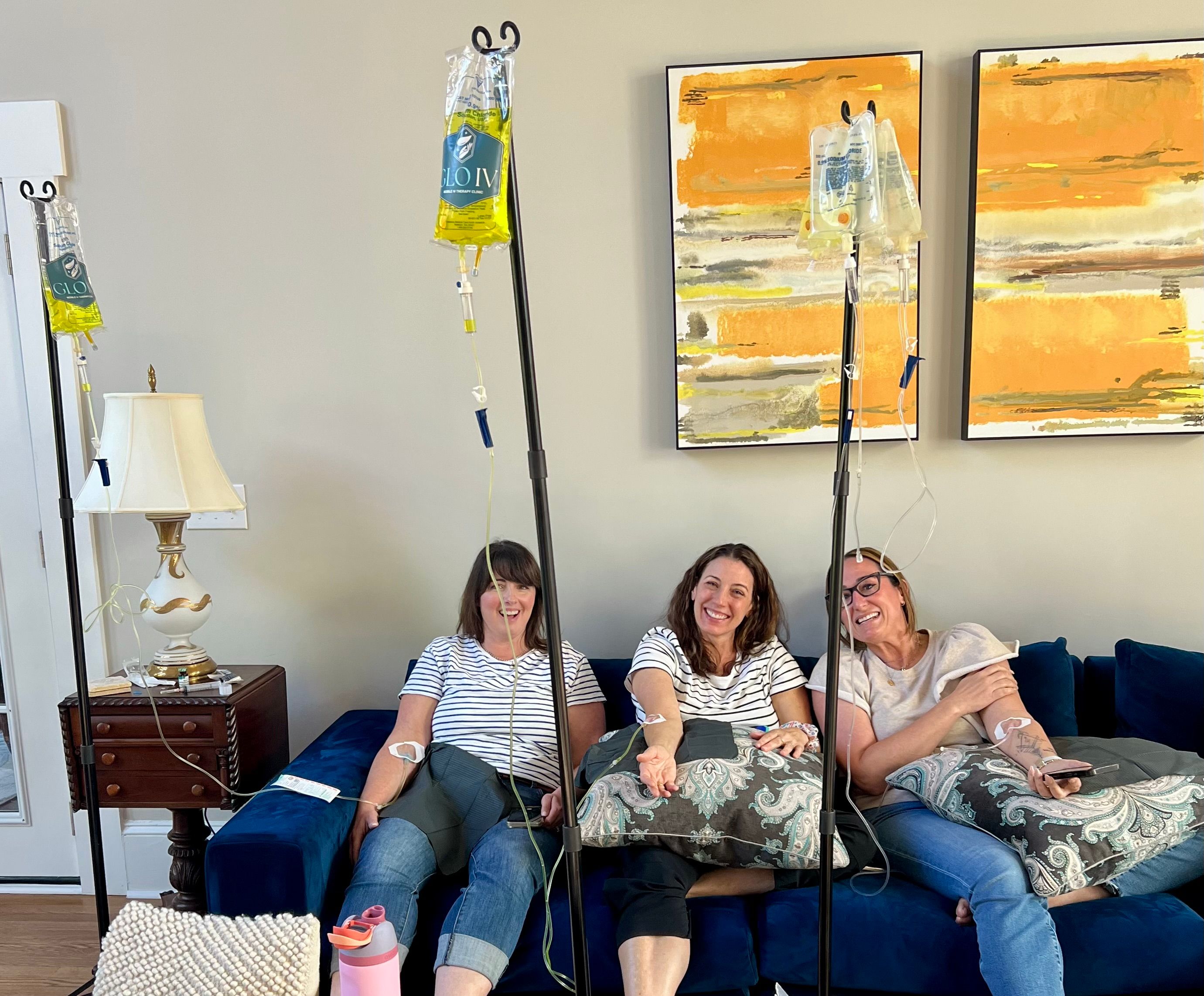Three women sitting on a blue couch in a cozy living-room style setting, smiling while receiving IV hydration drips from poles, with patterned pillows and orange abstract wall art behind them.
