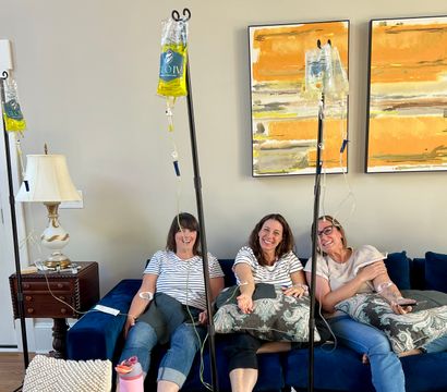 Three women sitting on a blue couch in a cozy living-room style setting, smiling while receiving IV hydration drips from poles, with patterned pillows and orange abstract wall art behind them.