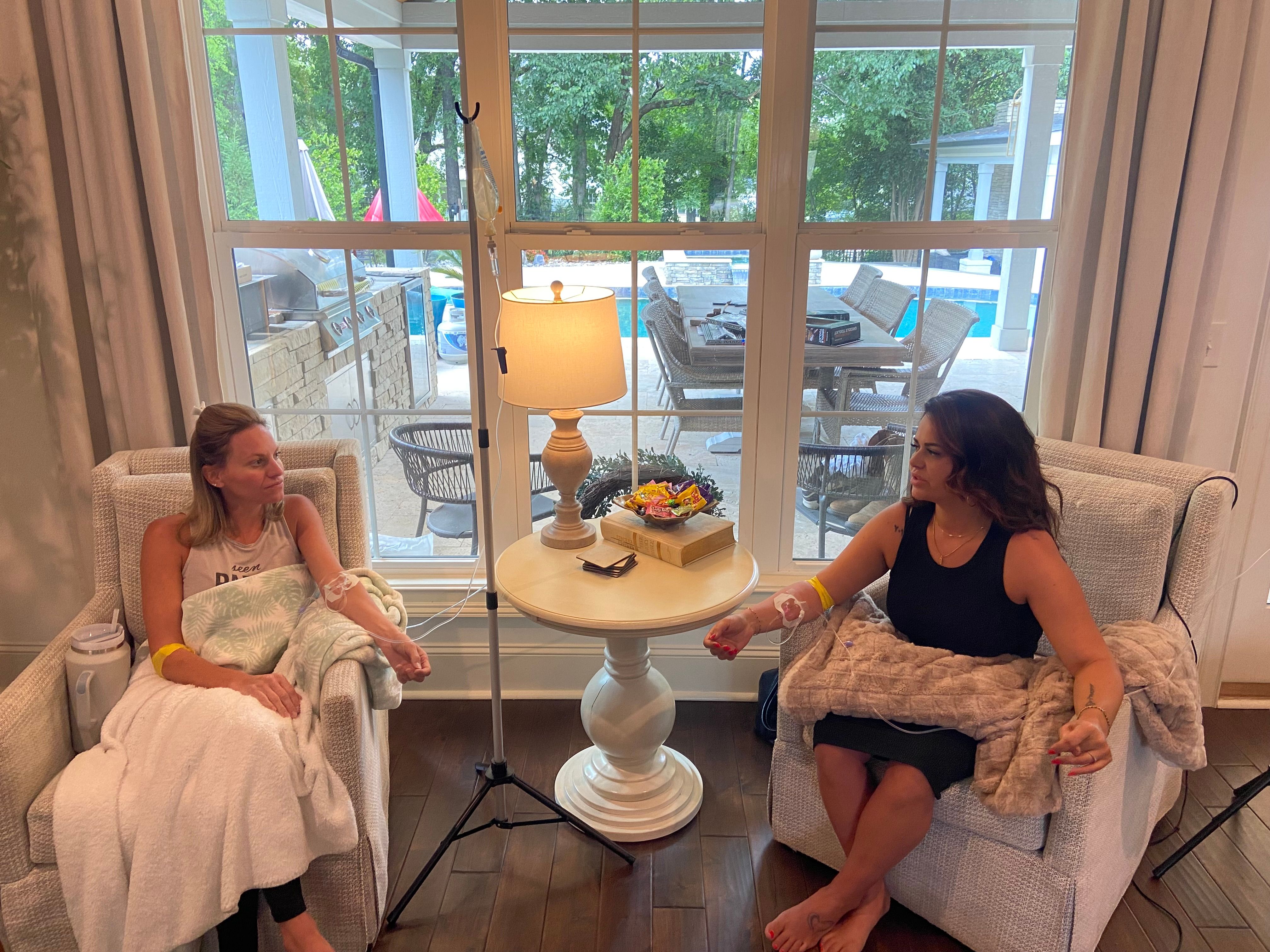Two people seated in armchairs receiving IV drips in a cozy living room, a round side table with lamp and snacks between them, and a large window showing a backyard patio and swimming pool outside.