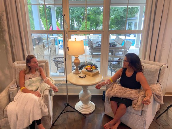 Two people seated in armchairs receiving IV drips in a cozy living room, a round side table with lamp and snacks between them, and a large window showing a backyard patio and swimming pool outside.