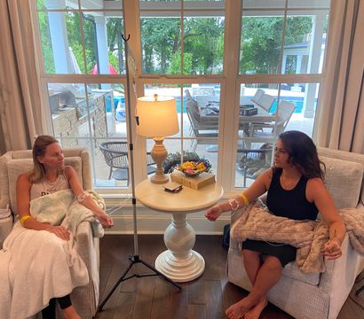 Two people seated in armchairs receiving IV drips in a cozy living room, a round side table with lamp and snacks between them, and a large window showing a backyard patio and swimming pool outside.