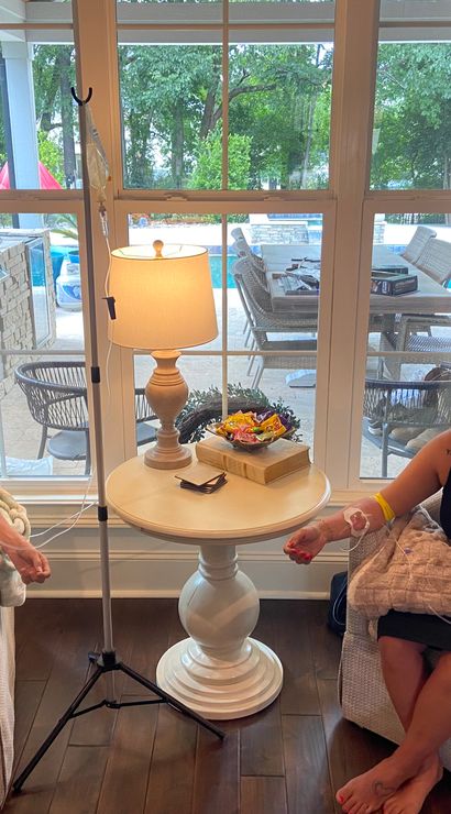 Two people seated in armchairs receiving IV drips in a cozy living room, a round side table with lamp and snacks between them, and a large window showing a backyard patio and swimming pool outside.