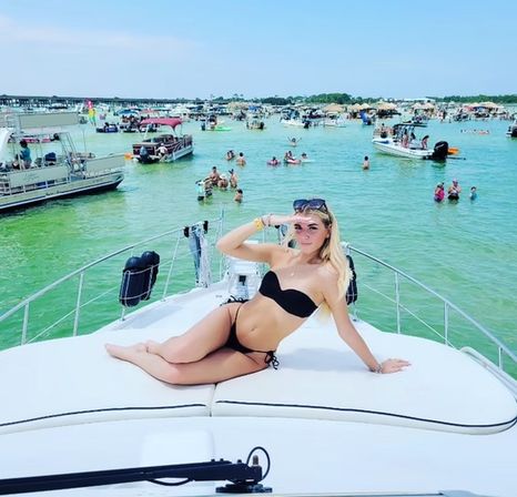 Person in a black bikini lounging on the bow of a white yacht at a busy sandbar with boats and people in shallow turquoise water on a sunny day