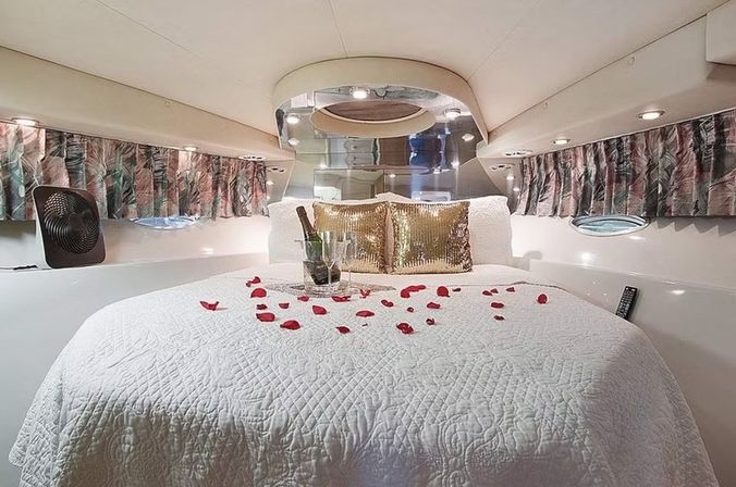 Romantic luxury yacht cabin bedroom with round white quilted bed, scattered red rose petals, champagne in an ice bucket and glasses, gold sequin pillows, porthole windows and soft recessed lighting.