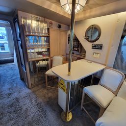 Cozy motor yacht salon bar with white countertop and brass pole, two padded bar stools, mirrored glassware shelves, round porthole, wood cabinets and plush carpet