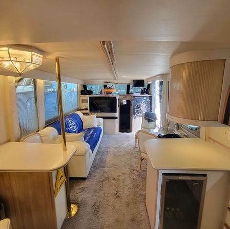 Cozy yacht interior salon with white leather sofa and blue throw pillow, carpeted floor, galley counters and mini-fridge, brass support pole and forward helm under warm overhead lighting.