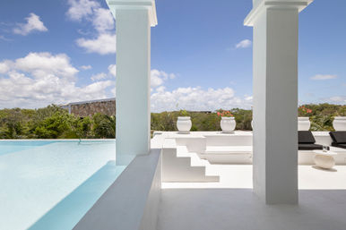 Sunlit white villa terrace with infinity pool framed by square columns, white planters with pink blooms, black lounge chairs and tropical greenery under a blue sky