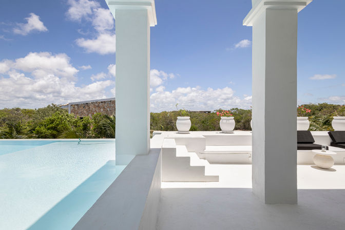 Sunlit white villa terrace with infinity pool framed by square columns, white planters with pink blooms, black lounge chairs and tropical greenery under a blue sky