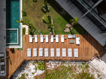 Aerial view of a coastal resort wooden sun deck with rows of white lounge chairs and side tables, turquoise pool and hot tub to the side, palm trees and green lawn, rocky beach below.