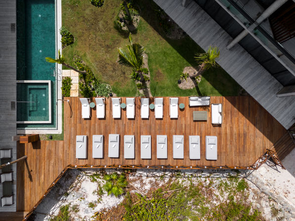 Aerial view of a coastal resort wooden sun deck with rows of white lounge chairs and side tables, turquoise pool and hot tub to the side, palm trees and green lawn, rocky beach below.