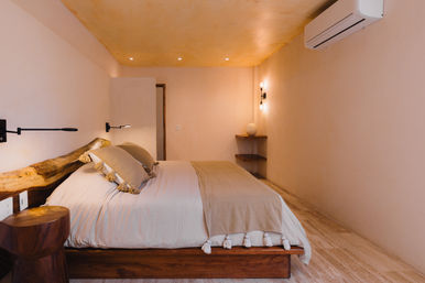 Cozy minimalist bedroom with low wooden platform bed, live-edge headboard, neutral linens and tassel throw, wall-mounted reading lamps, warm recessed and pendant lighting, small wooden stool and wall-mounted air conditioner.