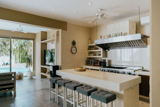 Sunlit open-concept modern kitchen with a large white island and five tufted gray bar stools, stainless-steel range hood and stove, and sliding glass doors opening to a pool and tropical plants.