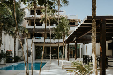 Tropical resort courtyard with blue swimming pool, white stucco building with thatched palapa roofs, palm trees, lounge chairs and shaded outdoor bar