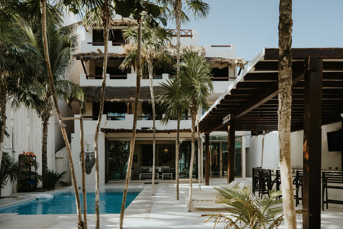 Tropical resort courtyard with blue swimming pool, white stucco building with thatched palapa roofs, palm trees, lounge chairs and shaded outdoor bar