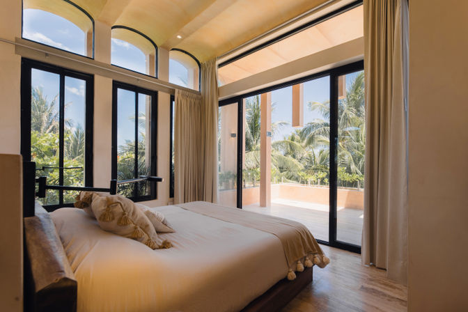 Sunlit modern bedroom in a coastal villa with a plush bed, floor-to-ceiling windows and sliding doors opening onto a tropical palm-lined balcony.