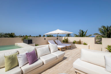 Sun-soaked rooftop terrace with white cushioned sofas and colorful pillows, small plunge pool, twin sun loungers under a white umbrella, tropical palms and blue ocean horizon
