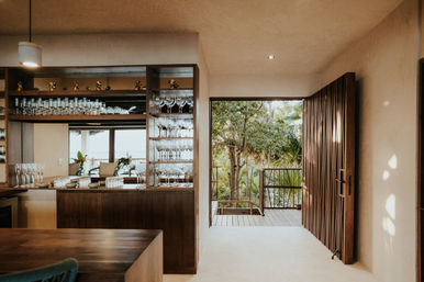 Sunlit tropical villa interior with wooden bar and shelves of wine glasses, open slatted door revealing a lush tree-lined balcony