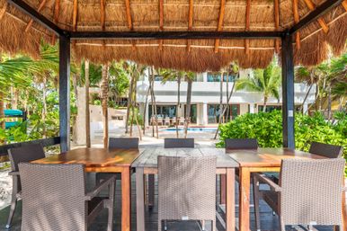 Sunny tropical palapa dining area with wooden table and wicker chairs overlooking a poolside white villa, palm trees and lush beachfront greenery.