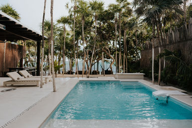 Tropical beachfront scene: turquoise rectangular pool with two lounge chairs under a wooden pergola, tall palms framing a sandy beach and ocean view beyond.