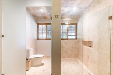 Bright spa-like modern minimalist bathroom with beige marble tiles, glass walk-in shower and rain showerhead, white toilet, wooden louvered window and recessed niche.