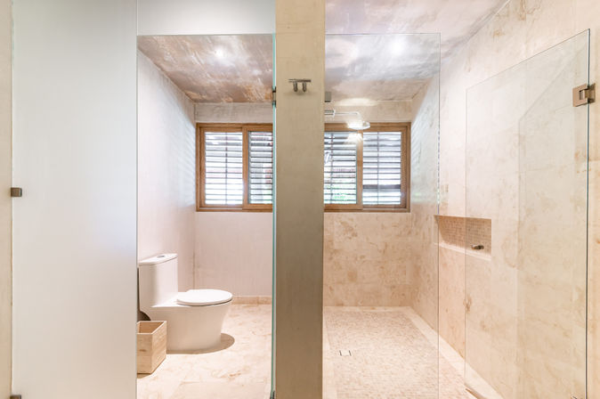 Bright spa-like modern minimalist bathroom with beige marble tiles, glass walk-in shower and rain showerhead, white toilet, wooden louvered window and recessed niche.