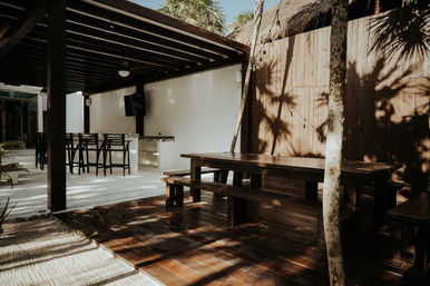 Sunlit tropical outdoor patio with a dark wooden picnic table and benches on a deck, bar stools under a shaded pergola, palm tree shadows and hanging string lights.