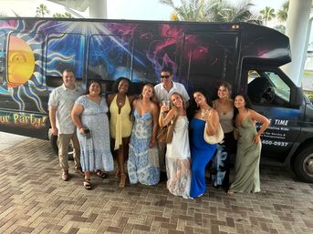 Smiling group of friends in summer dresses and casual shirts posing in front of a colorful cosmic-themed party shuttle under palm trees, holding drinks and ready to celebrate