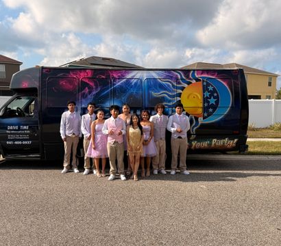 Prom group of teens in pastel formalwear posing in front of a colorful sun-and-moon party bus parked on a suburban street under a cloudy sky